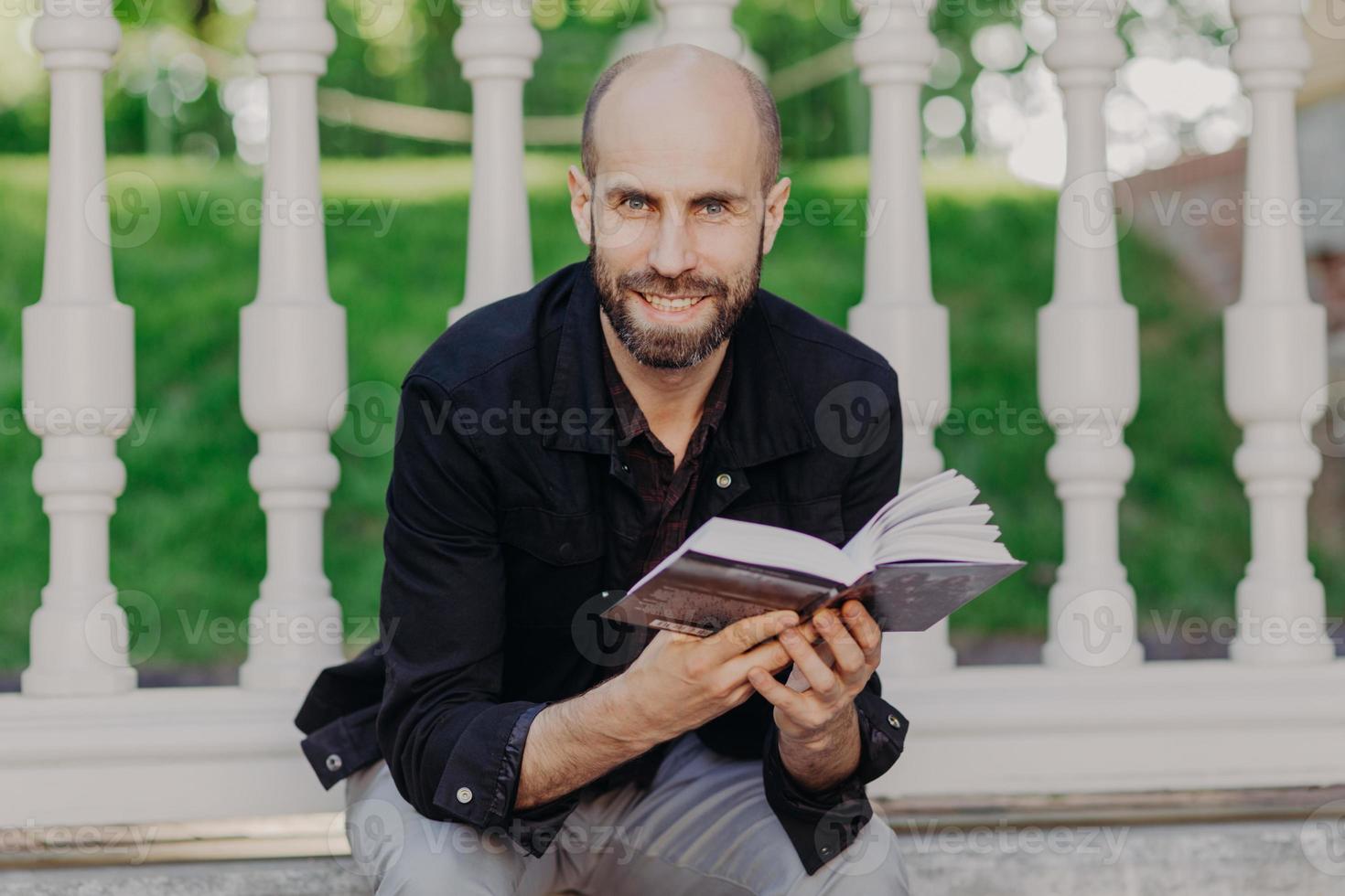 homem barbudo positivo olha diretamente para a câmera, lê um livro muito emocionante e interessante, senta-se contra o fundo da varanda, passa o tempo livre no jardim de verão. pessoas, educação e felicidade foto