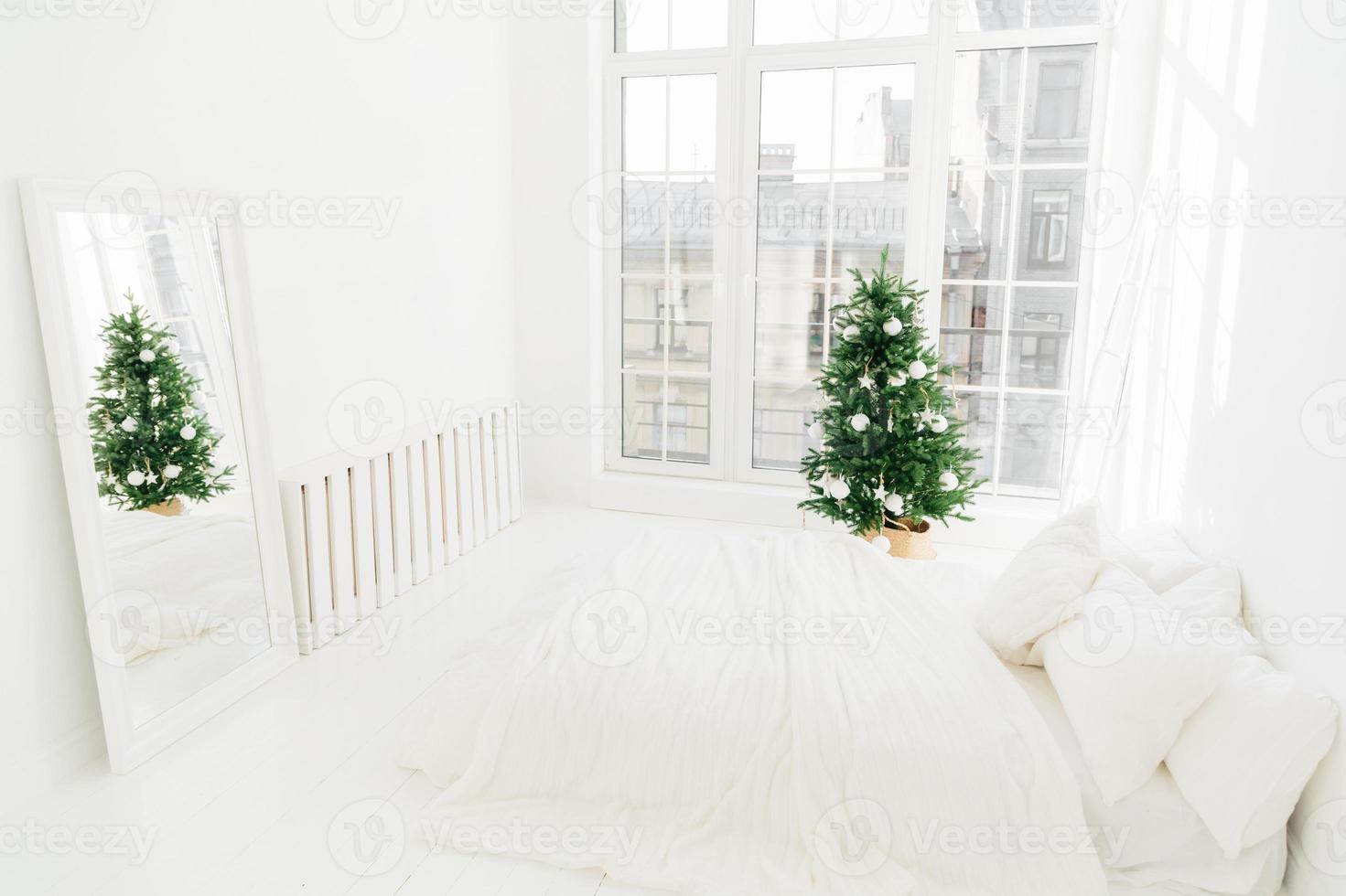 conforto, lar e conceito de véspera de natal. quarto com cama macia branca, espelho, grande janela para luz entrando no quarto, árvore de ano novo decorada e escada. decoração de férias. foto