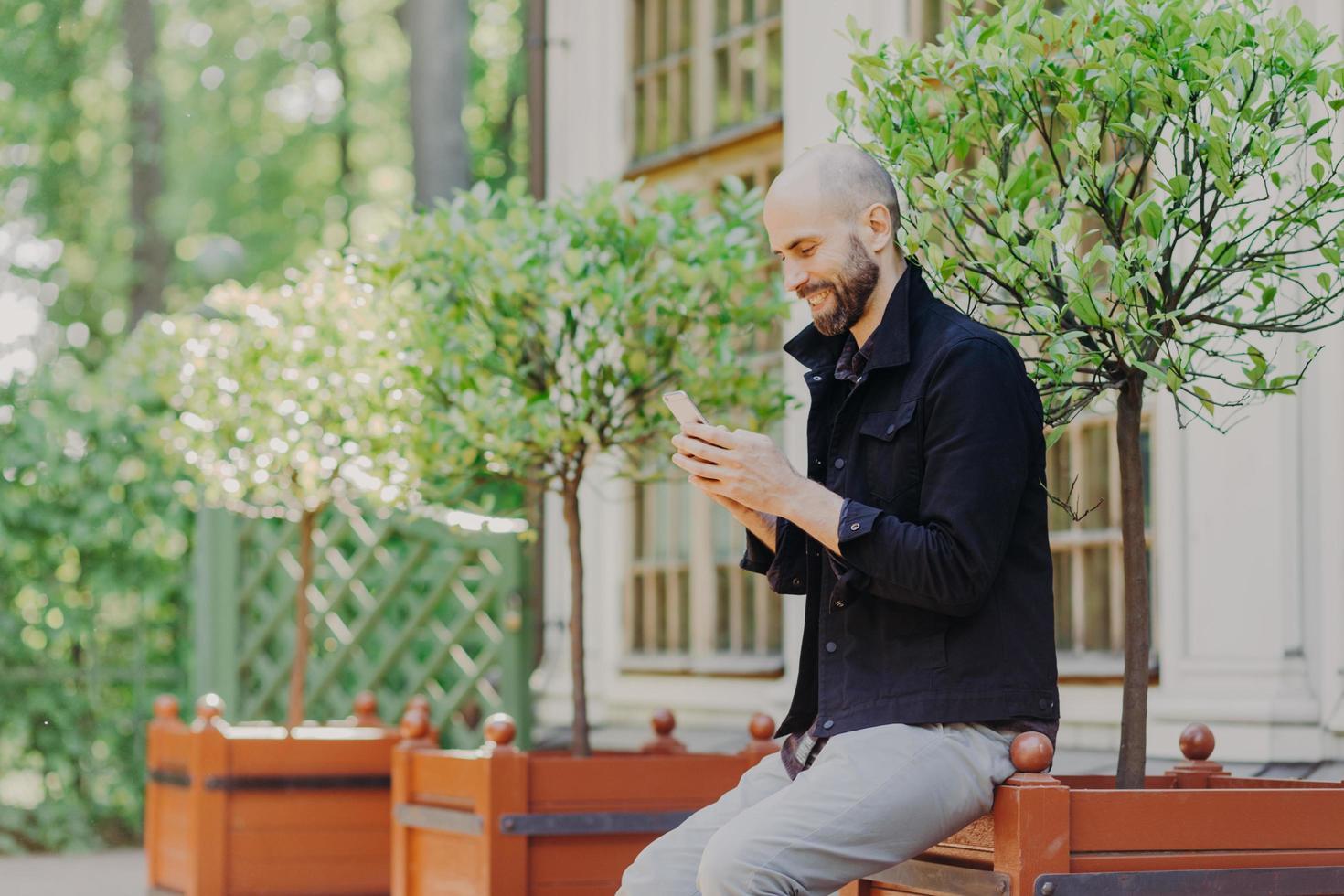 freelancer masculino concentrado gosta de dia de folga, respira ar fresco ao ar livre, usa celular moderno, vê fotos em redes sociais tem sorriso positivo no rosto. cara alegre digita mensagem de texto no celular