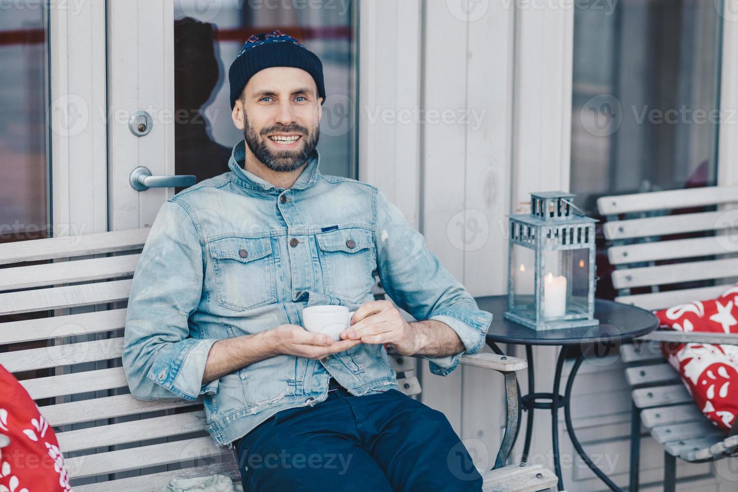 conceito de pessoas, descanso e positividade. sorrindo encantado modelo masculino barbudo com expressão feliz descansa no café aconchegante, segura caneca com chá, estando em alto astral, pensa em algo agradável foto