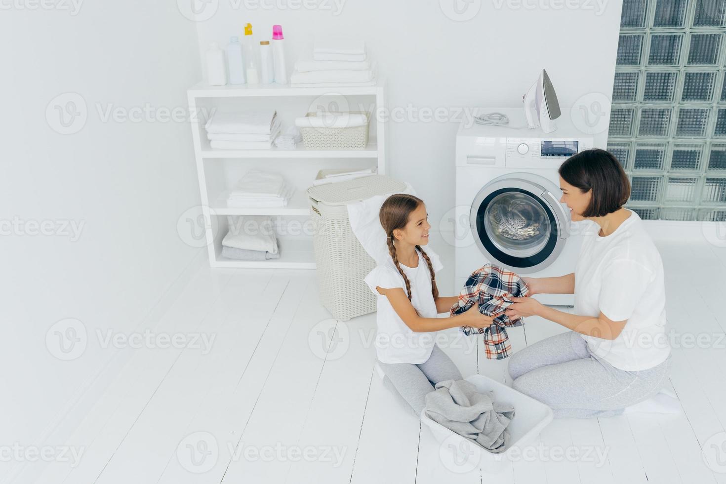 mãe dona de casa e filha carregam a máquina de lavar com roupas sujas, posam na espaçosa lavanderia, fazem tarefas em casa, olham felizes uma para a outra. conceito de dia de lavanderia. família na sala de lavagem foto