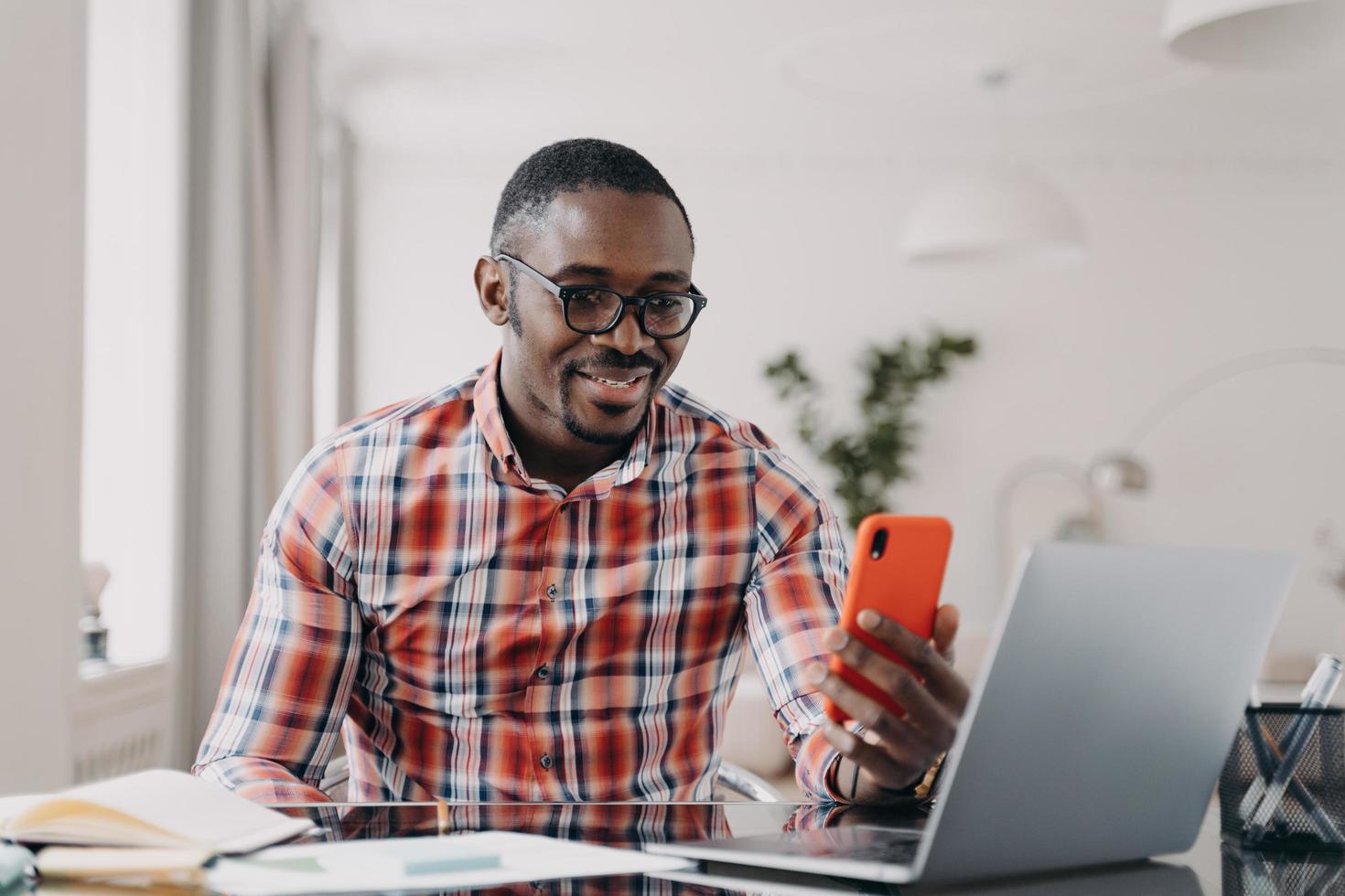 homem afro feliz tem chamada de vídeo no telefone. cara fica em casa em quarentena e trabalhando remotamente no laptop. foto