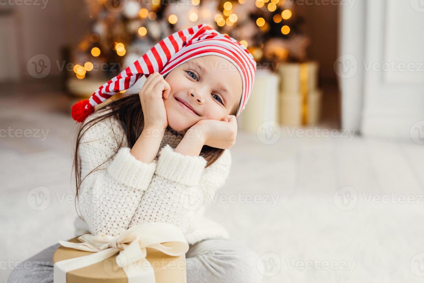 linda criança pequena posa na sala de estar, inclina-se no presente presente, tem expressão feliz, feliz em receber surpresa dos pais, passa férias no círculo familiar. feliz Natal e Feliz Ano Novo foto