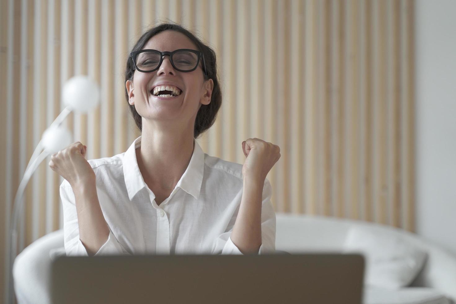 Senhora italiana animada cerrando os punhos com alegria enquanto está sentado na frente do laptop no local de trabalho foto