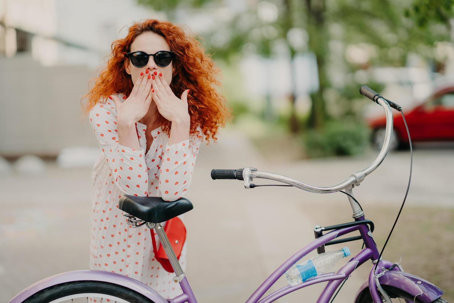 foto ao ar livre de linda mulher ruiva cobre a boca com as duas palmas, tem manicure vermelha, vestida com vestido da moda e óculos de sol da moda, posa perto de sua bicicleta, passa o tempo livre andando de bicicleta