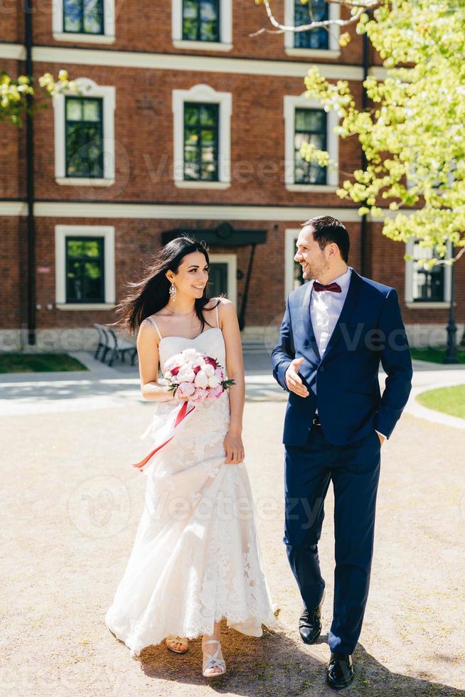 linda jovem noiva no vestido de noiva branco, segura o buquê, conversa com o futuro marido, caminha juntos, vai ao cartório. lindo casal se alegra dia especial em sua vida. foto