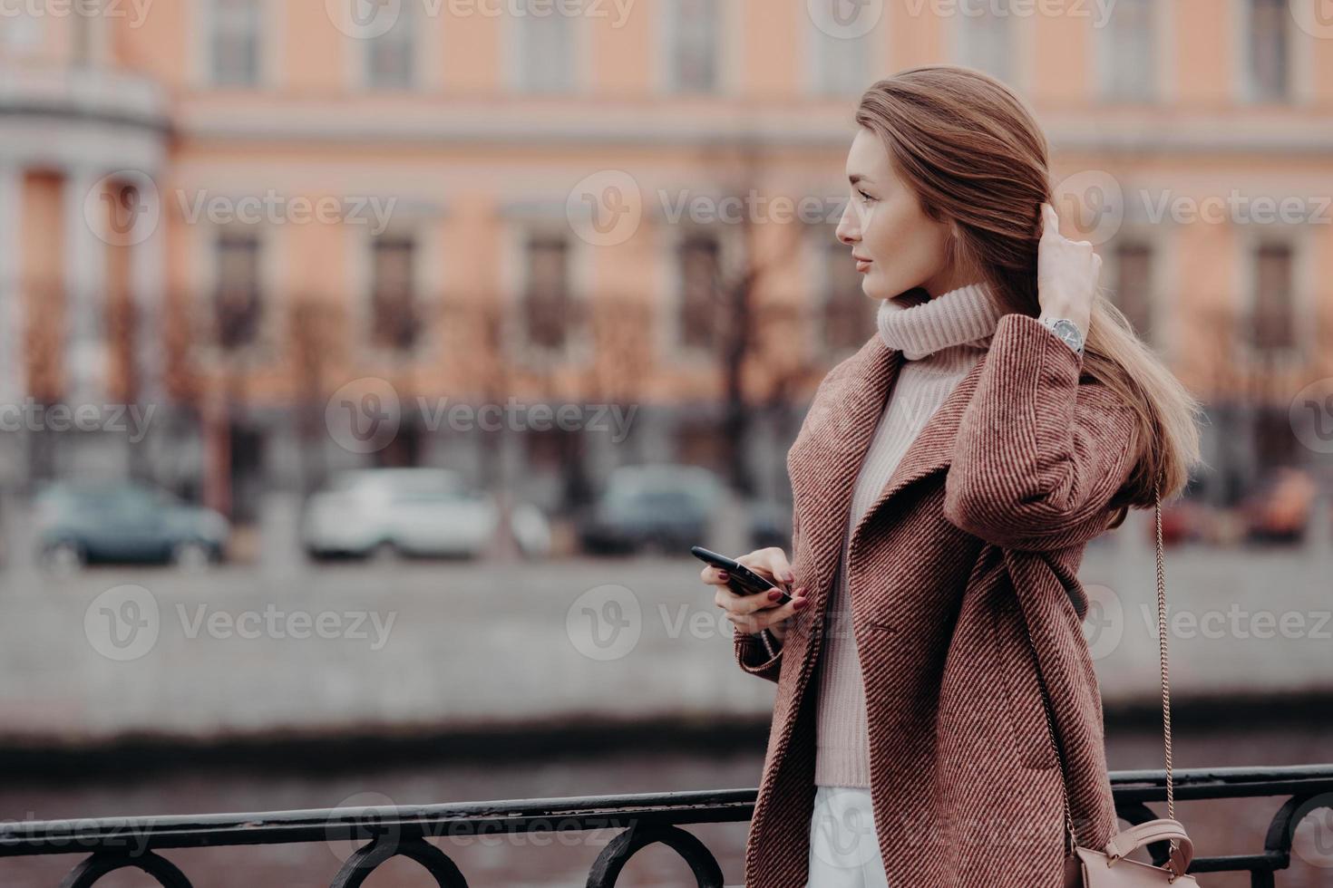 foto de modelo feminino jovem e atraente com cabelo comprido, vestido com casaco, tem passeio ao ar livre durante o outono na cidade grande, segura telefone celular moderno, mensagens de texto, admira a bela paisagem urbana