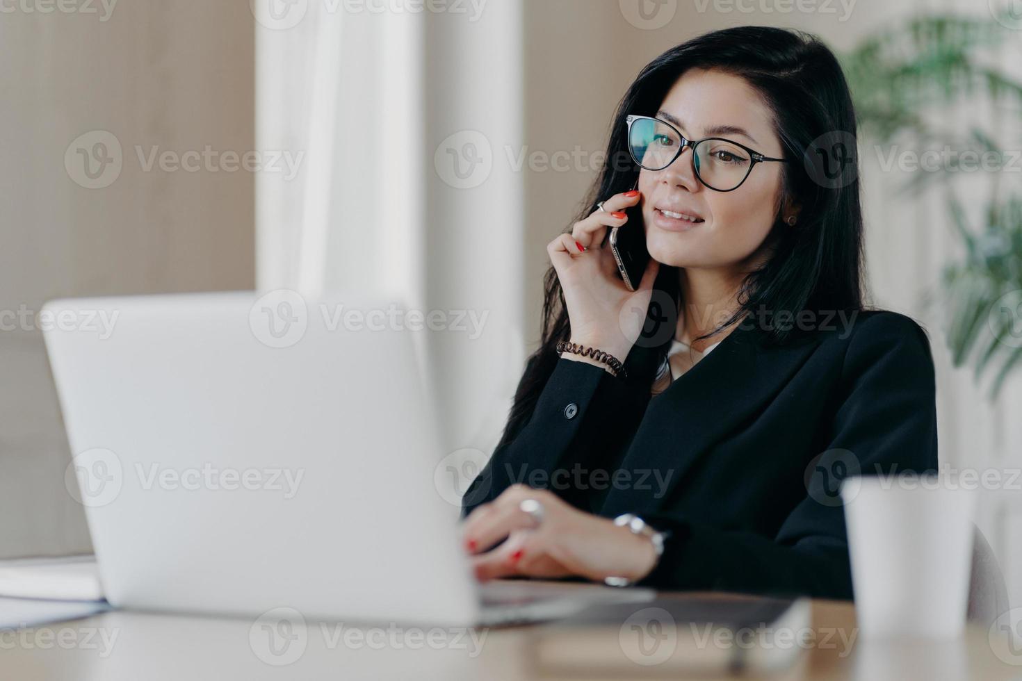 mulher de negócios séria confiante tem conversa telefônica, posa na frente do computador portátil, usa óculos ópticos para correção da visão, posa no local de trabalho, faz trabalho de pesquisa, vestida formalmente foto