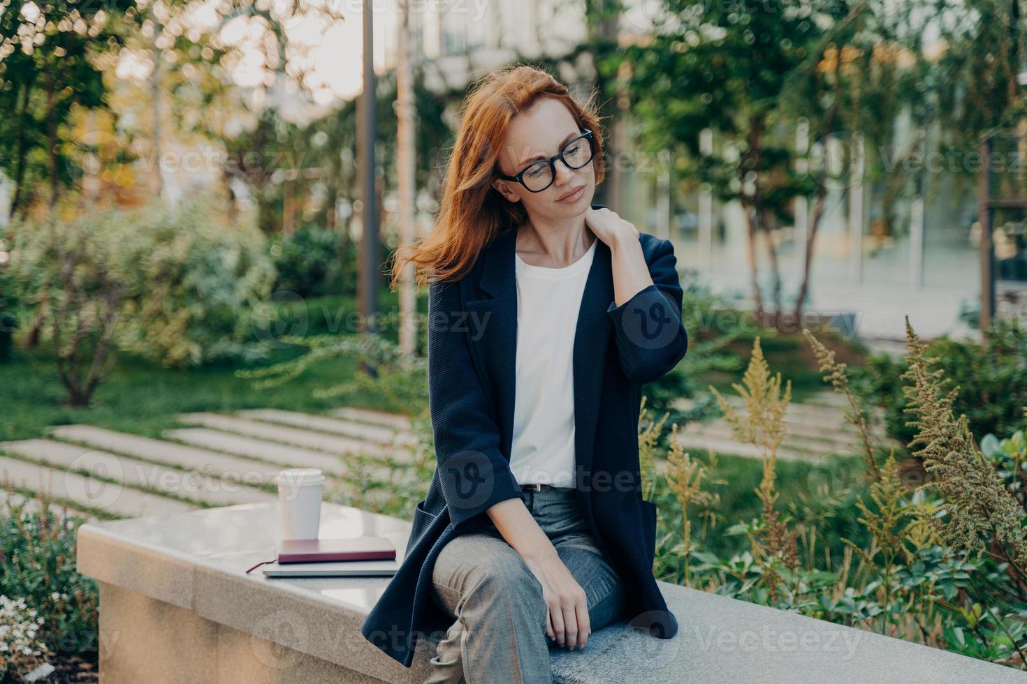 mulher ruiva cansada descansa no parque sofre de dor no pescoço se senta no banco foto