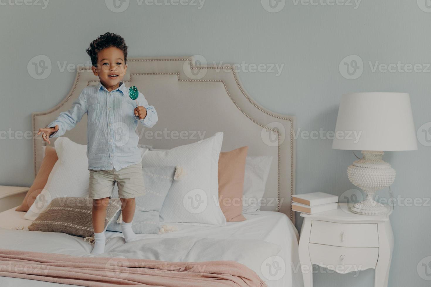 menino de criança afro-americana bonito despreocupado com pirulito pulando ob cama em casa foto