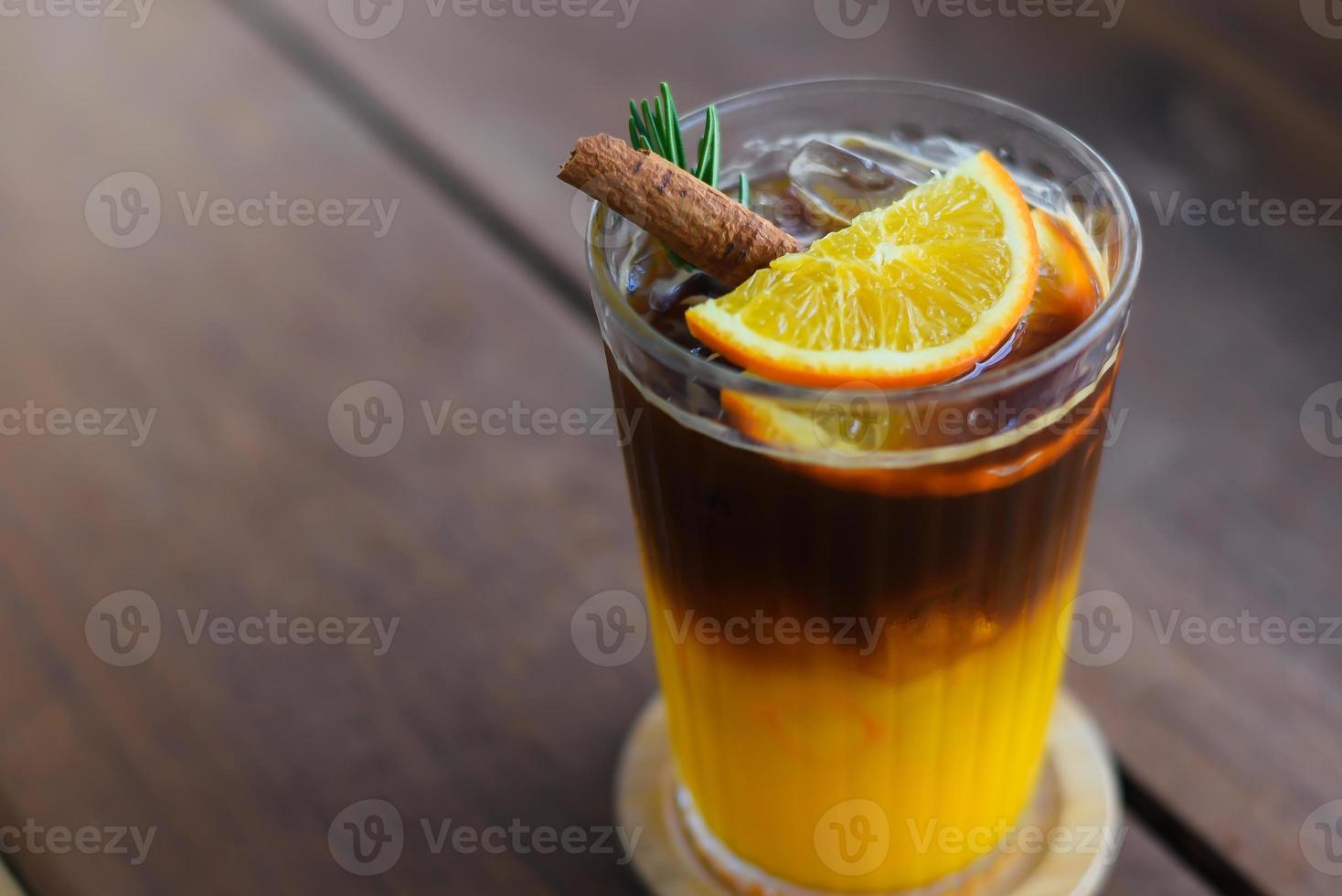 café preto gelado misturado com suco de laranja em cobertura de vidro com laranjas fatiadas, folhas de alecrim e canela por cima. em uma montanha-russa de madeira colocada sobre uma mesa de madeira. foto