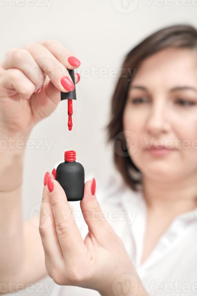 mulher morena segurando o esmalte vermelho aberto e mostrando o pincel. vista de corte. manicure e cuidados com as unhas. cor brilhante para férias. manicure feminina na moda elegante. foto