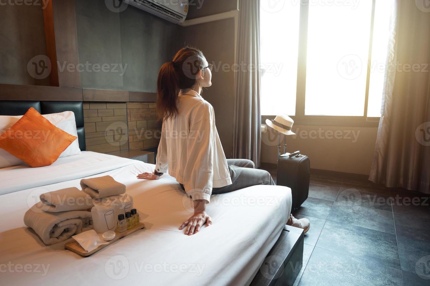 mulher turista sentada na cama com sua bagagem no quarto do hotel após o check-in e olhando para uma bela vista do lado de fora da janela. conceitual de viagem e hospedagem. foto