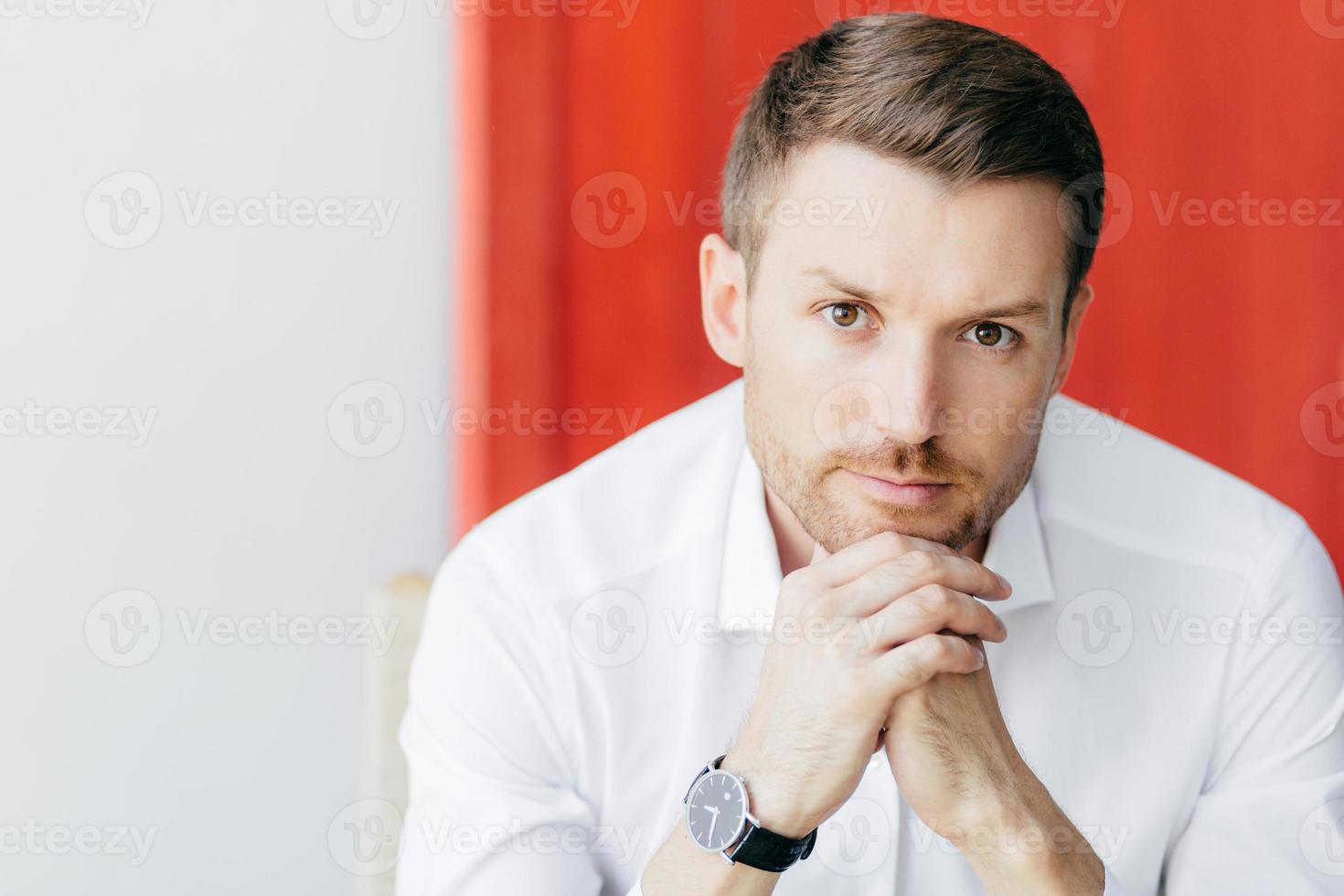 tiro de homem com barba pensativo atraente segura o queixo, vestido com camisa branca elegante, espera por alguém, sendo empresário próspero, posa contra fundo branco e vermelho com espaço livre à parte foto