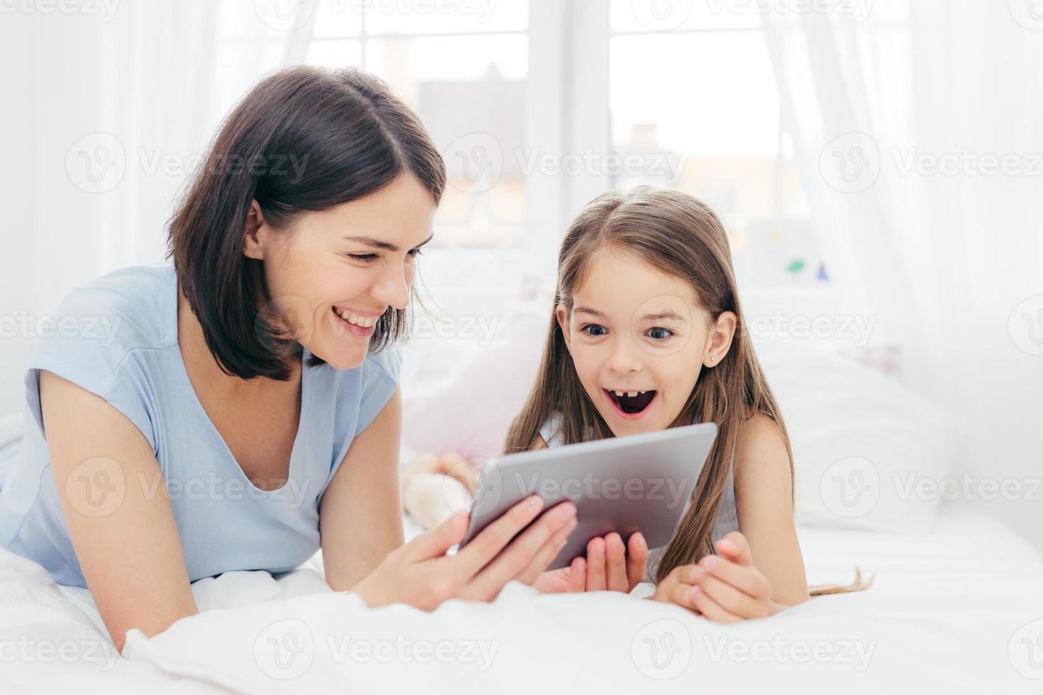 linda mãe e filha assistem a algo engraçado no computador tablet, conectado à internet sem fio, passam o tempo livre no quarto, surpreendem a expressão feliz. conceito de pessoas e lazer foto
