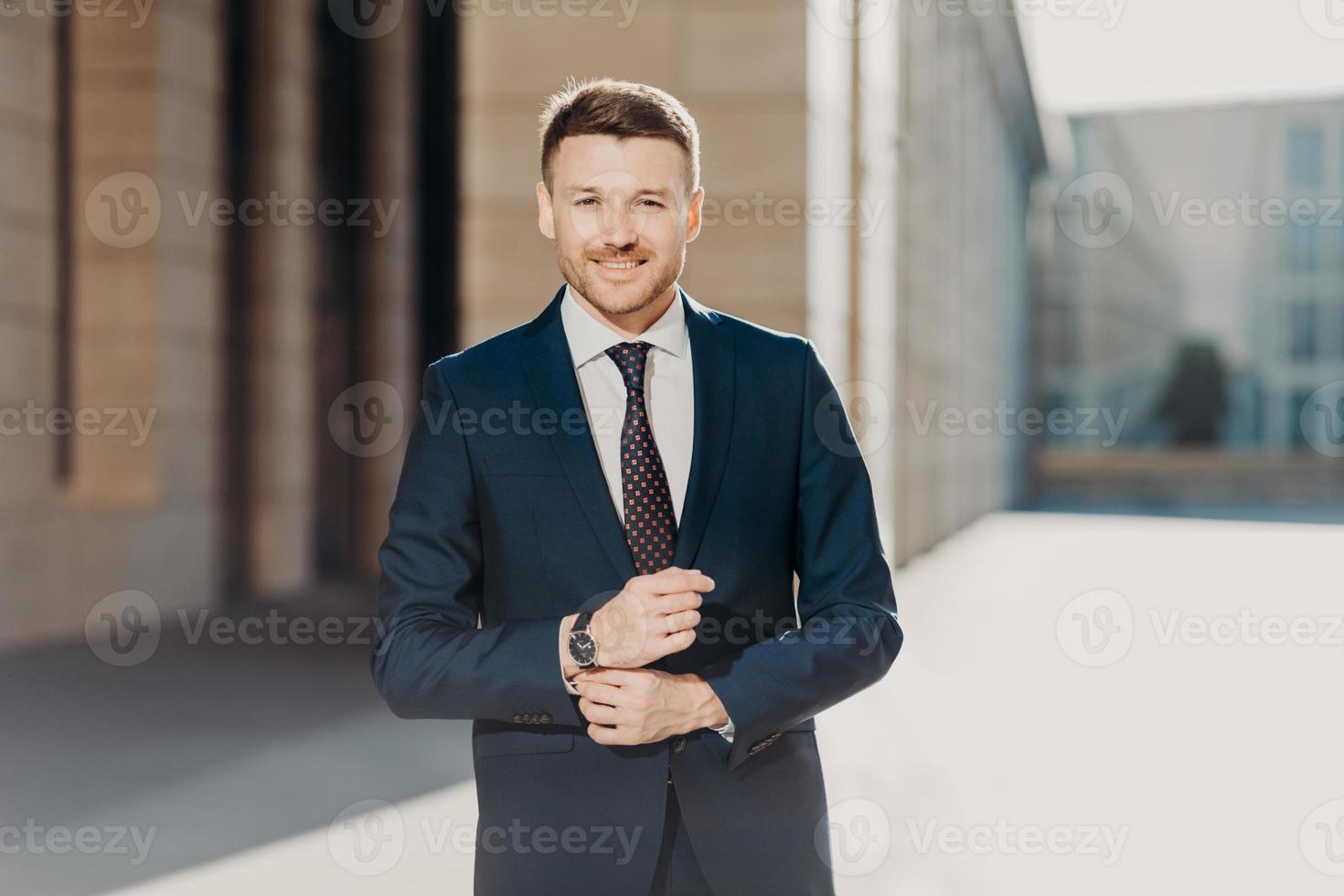 empresário bem sucedido com expressão alegre, vestido com roupas formais, fica ao ar livre perto de sua empresa, indo se encontrar com colegas, olha diretamente para a câmera. pessoas, ocupação e negócios foto