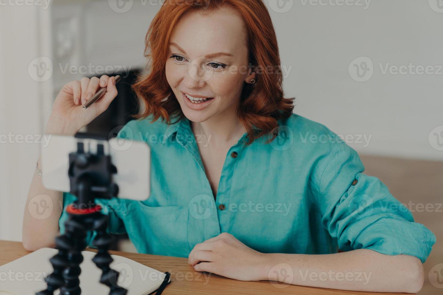 jovem profissional vlogger ou blogueira posa na frente da câmera do smartphone, dá conselhos sobre como iniciar o próprio negócio, senta-se na área de trabalho, segura a caneta. pessoas, tecnologia, estilo de vida, conceito de blogs foto