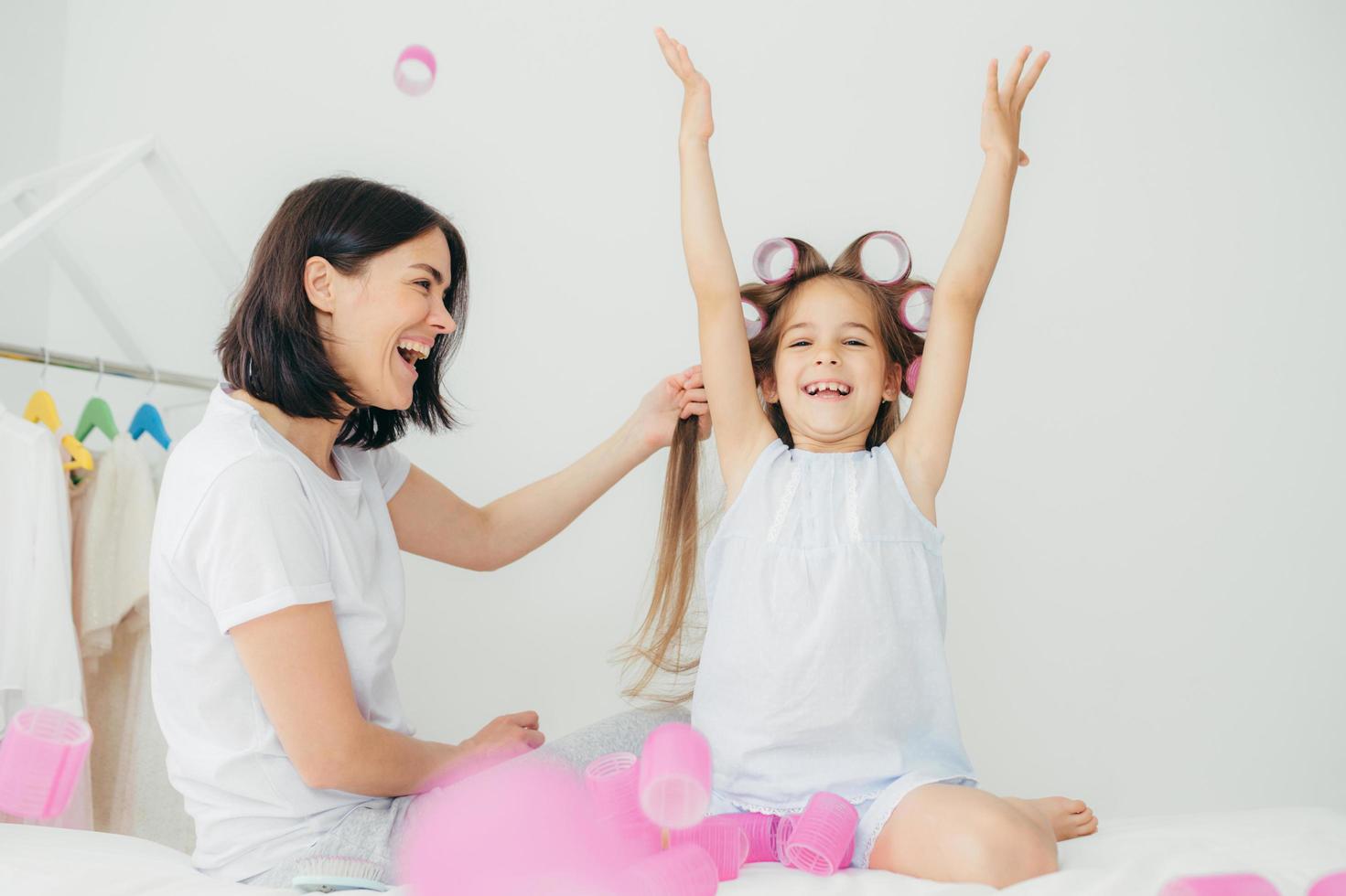 alegre mulher morena faz o cabelo de sua filha que tem expressão feliz, levantou as mãos e tem rolinhos na cabeça, sorri com prazer. família amigável se diverte junto, faz tratamentos de beleza. foto