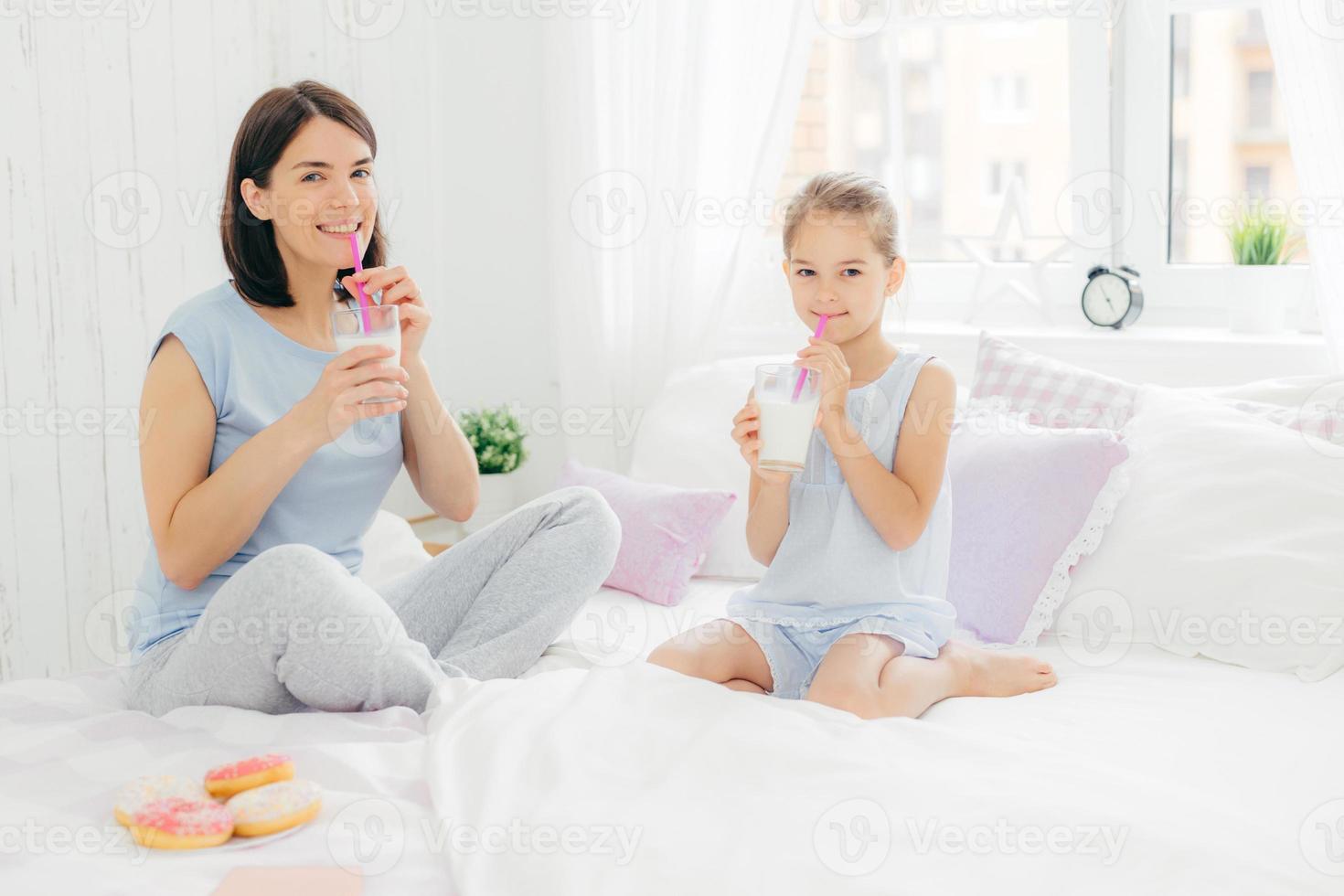 alegre mãe e filha vestidas de pijama, tomam café da manhã, bebem milk-shake com rosquinhas, sentam as pernas cruzadas na cama confortável no quarto branco. pessoas e conceito de estilo de vida foto