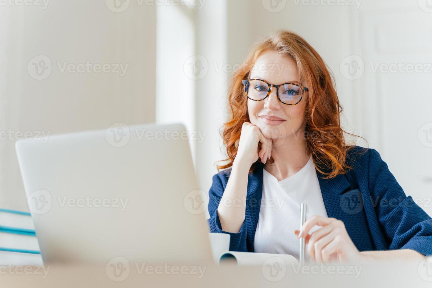 tiro horizontal de advogada profissional bem-sucedida de aparência agradável aprende caso de clientes, trabalha em computador portátil moderno, vestido com roupas formais e óculos transparentes, posa no escritório foto