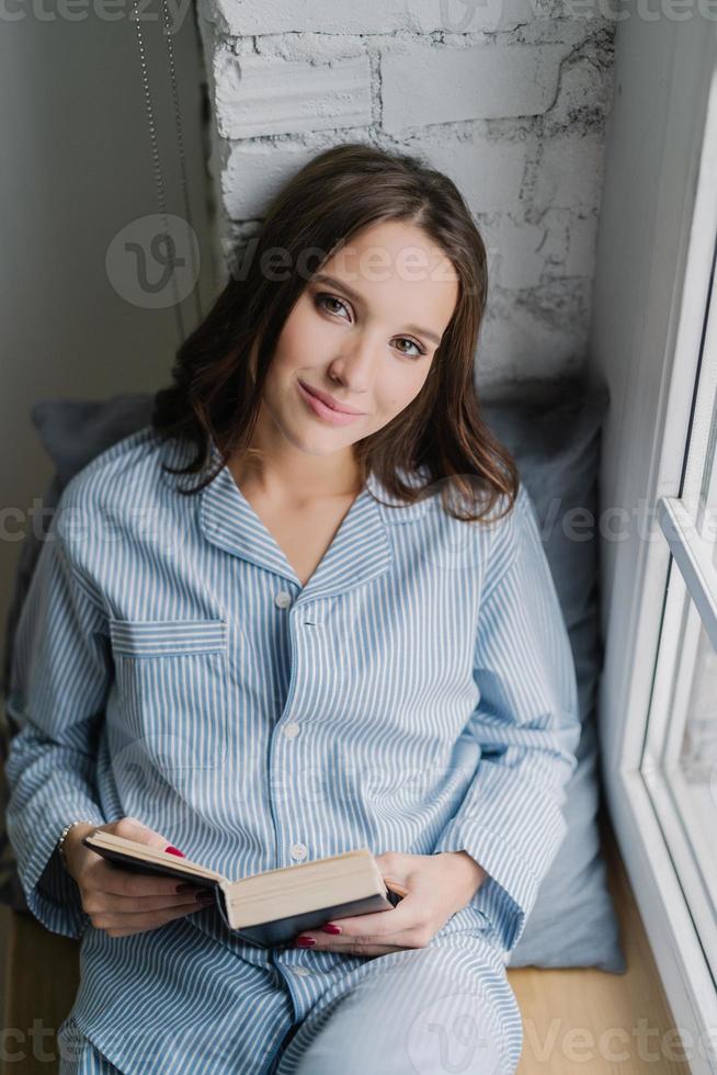 linda mulher relaxada com expressão despreocupada, vestida com roupa de dormir, lê livro no peitoril da janela de manhã, gosta de tempo livre, gosta de seu hobby. tiro vertical de adolescente com literatura foto