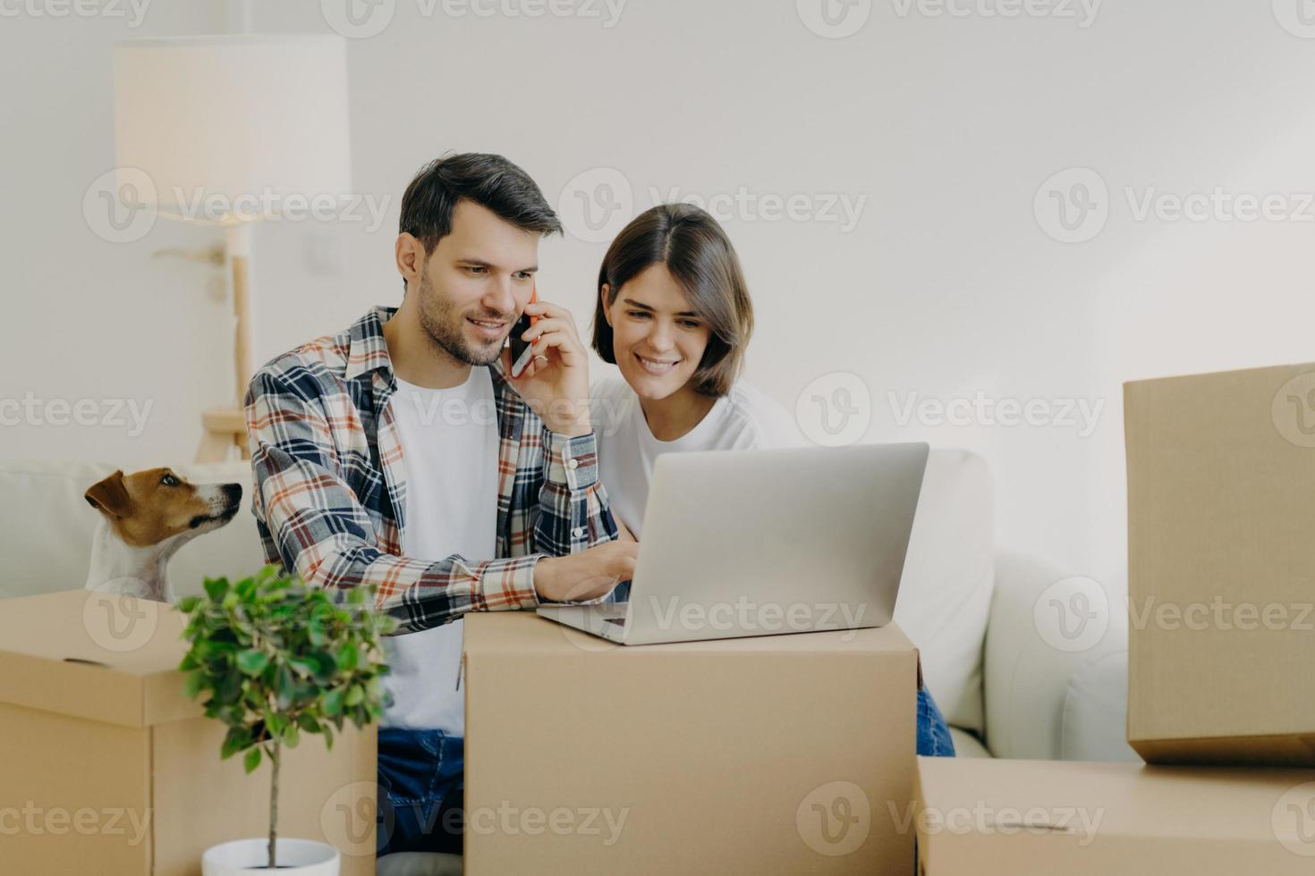 homem feliz navega no laptop em um novo apartamento, liga via smartphone, muda-se para um novo apartamento junto com a esposa, senta-se no sofá confortável, cercado de caixas de papelão na sala de estar, cachorro pequeno perto. foto