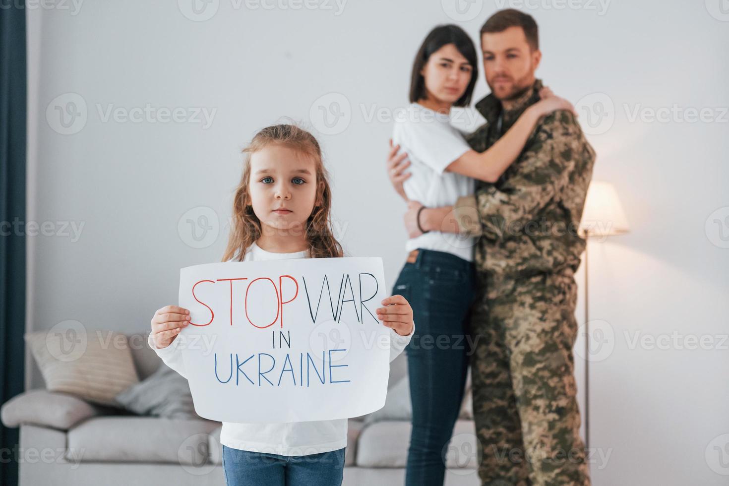garota segurando o banner com texto de parar a guerra nele. soldado de uniforme está em casa com sua esposa e filha foto