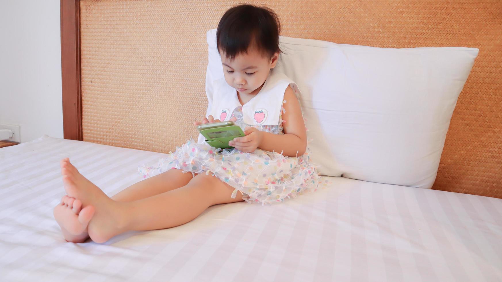 retrato de encantadora menina asiática de 3 anos senta-se na cama joga o jogo no smartphone. foto