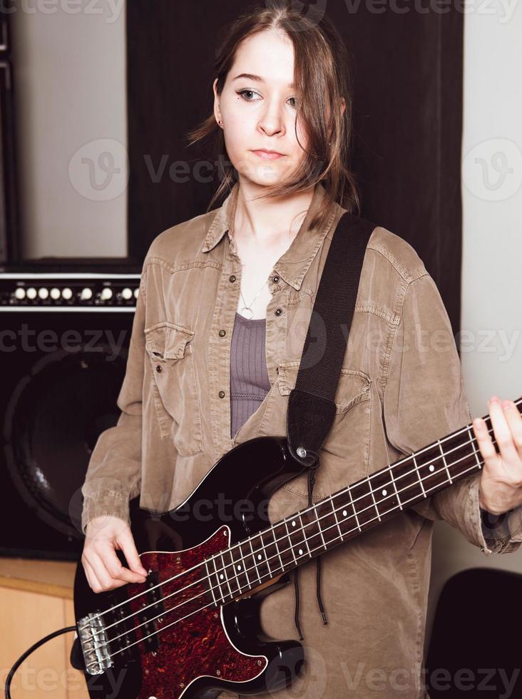 jovem vestindo roupas casuais e tocando baixo elétrico olhando de lado. baixista feminino em pé em uma sala de ensaio foto