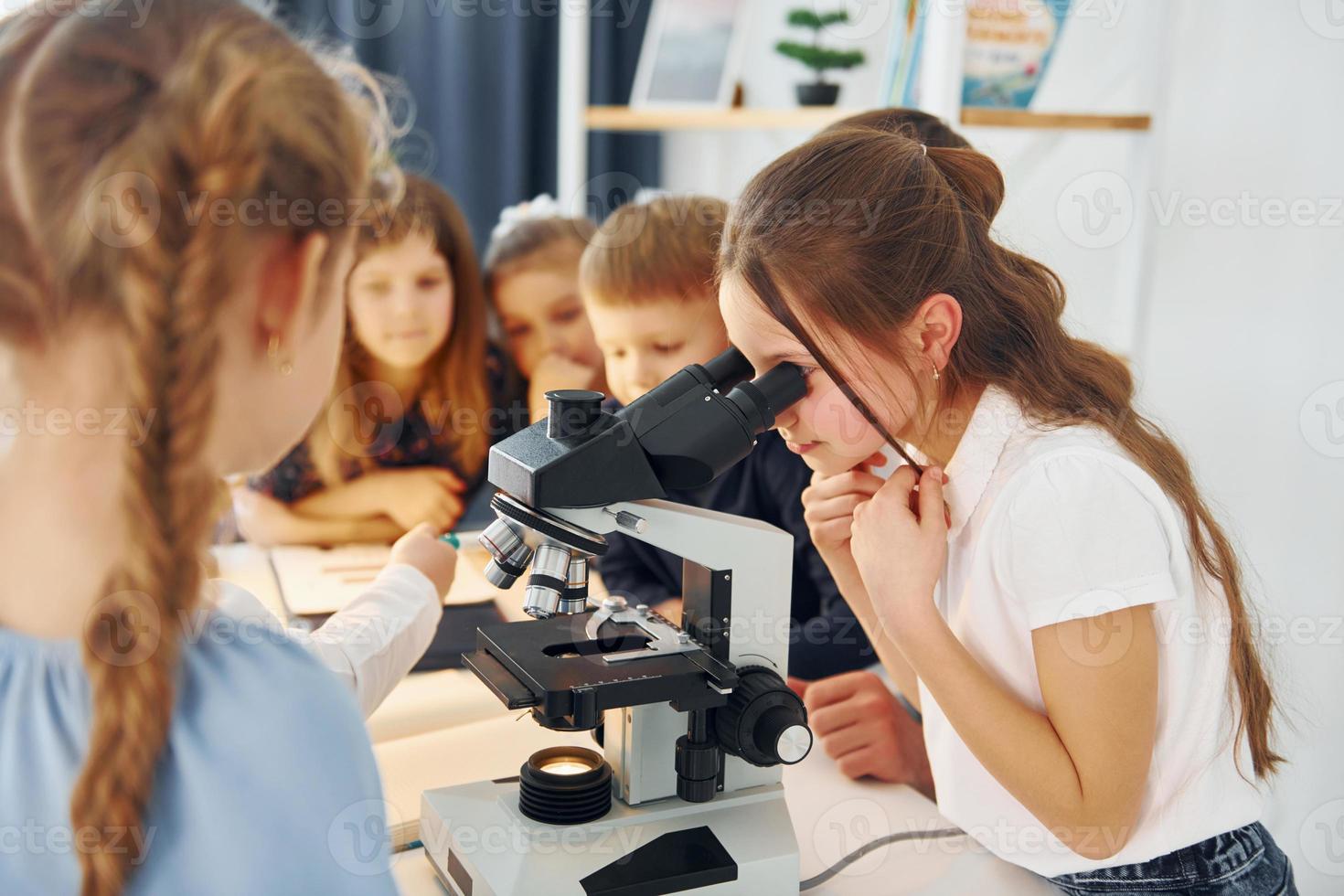 garota olhando no microscópio. grupo de alunos de crianças em sala de aula na escola com professor foto