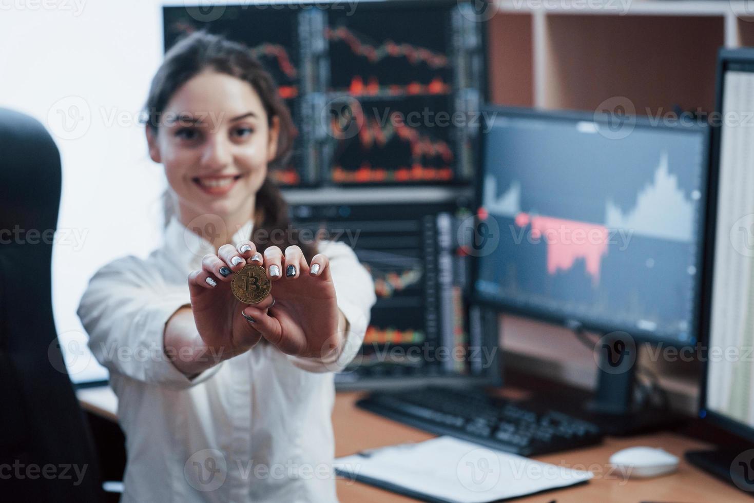 humor alegre. linda mulher segurando bitcoin nas mãos enquanto está sentado no escritório moderno com muitos monitores com gráficos foto