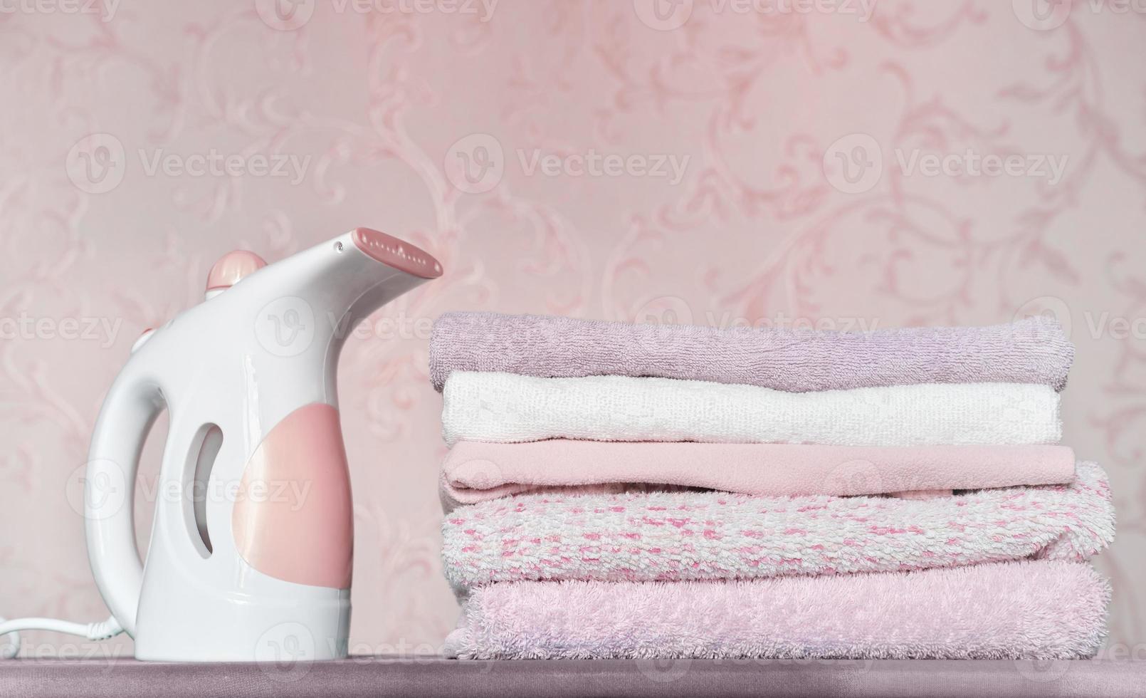 vaporizador de roupa elétrico portátil e pilha de toalhas dobradas na tábua de passar roupa. limpeza e agregado familiar, conceito de tarefas. roupas rosa e lilás. foto