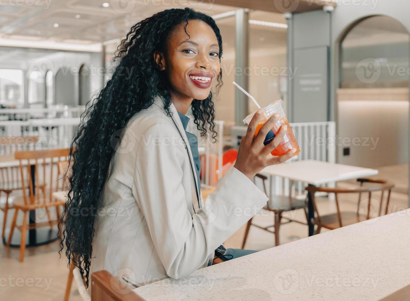 retrato de mulher negra com estilo de cabelo afro longo no café bar, bebendo bebida gelada, olhando para a câmera sorrindo. empresária gosta de relaxar com um copo de cafeína na pausa para o café para se refrescar. foto