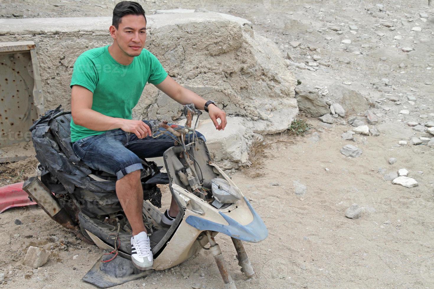 um jovem de cabelo escuro senta-se em uma scooter destruída em um estacionamento em mykonos, grécia. foto