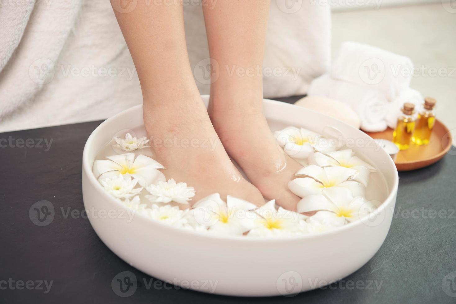 closeup vista de mulher encharcando os pés no prato com água e flores no piso de madeira. tratamento de spa e produto para pés femininos e spa de mãos. flores brancas em tigela de cerâmica. foto