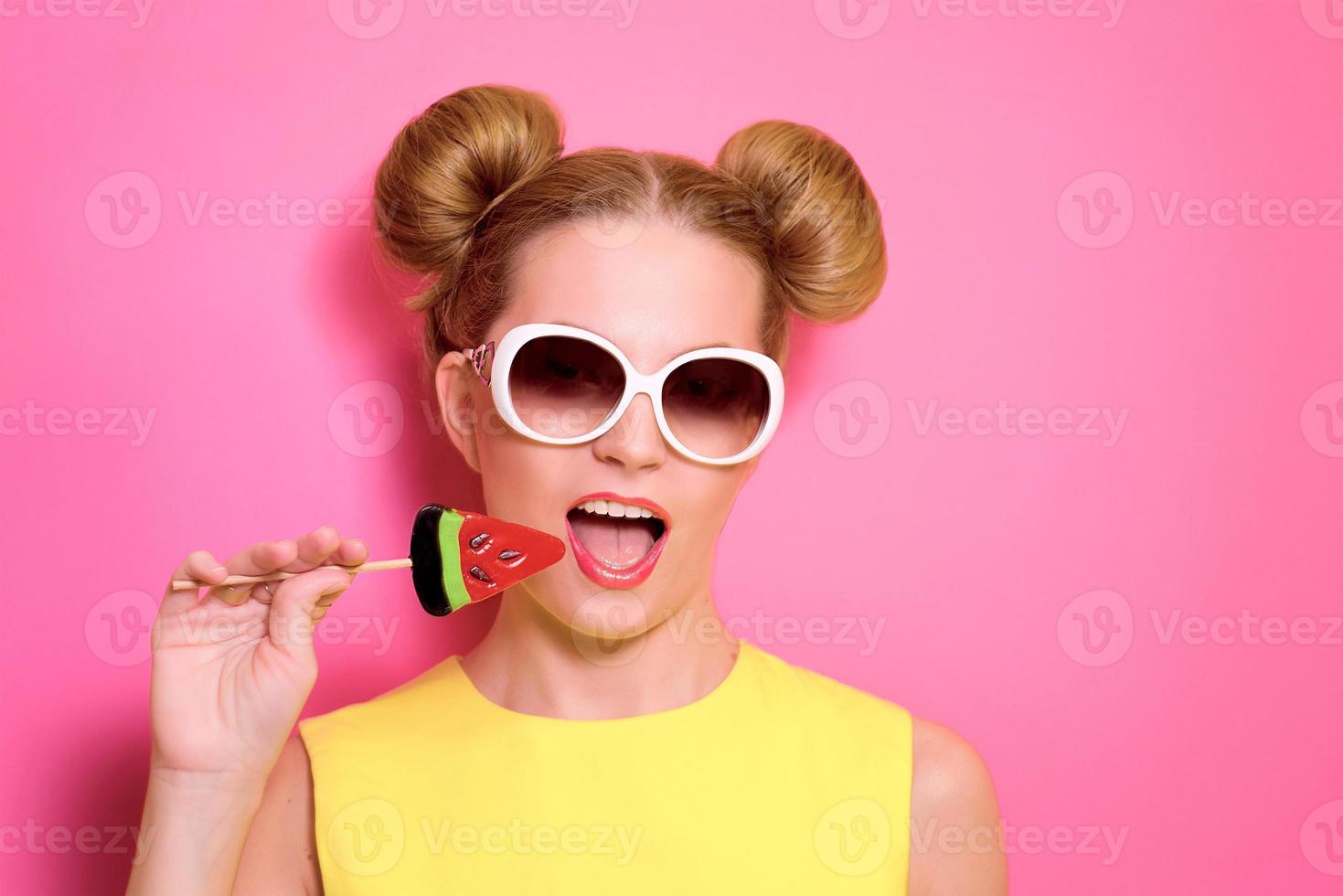 retrato de uma jovem loira bonita elegante em óculos de sol com pirulito de melancia colorido no fundo rosa foto