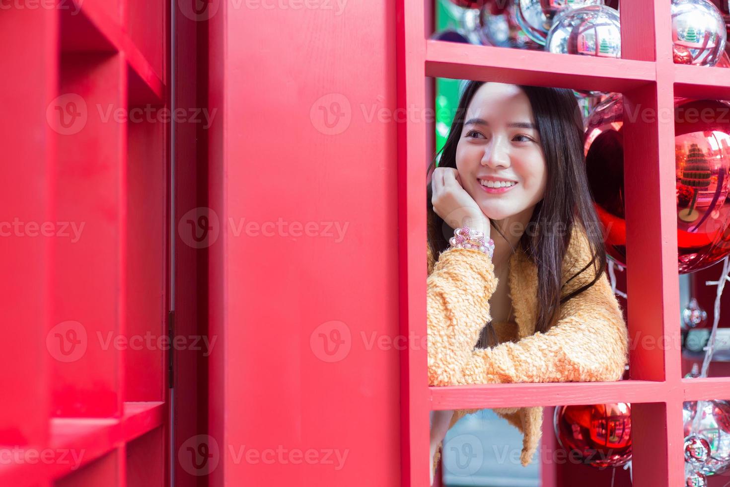 mulher bonita asiática de cabelos compridos vestindo um manto amarelo e sorriso feliz em pé na cabine telefônica vermelha no tema de celebrar o natal e feliz ano novo foto