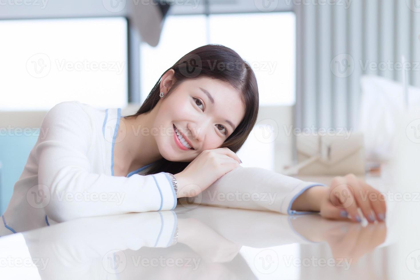 retrato de mulher jovem e bonita alegre está se sentindo feliz e sorrindo deitado na mesa. modelo de mulher sente-se na sala envidraçada como sala de negócios. foto