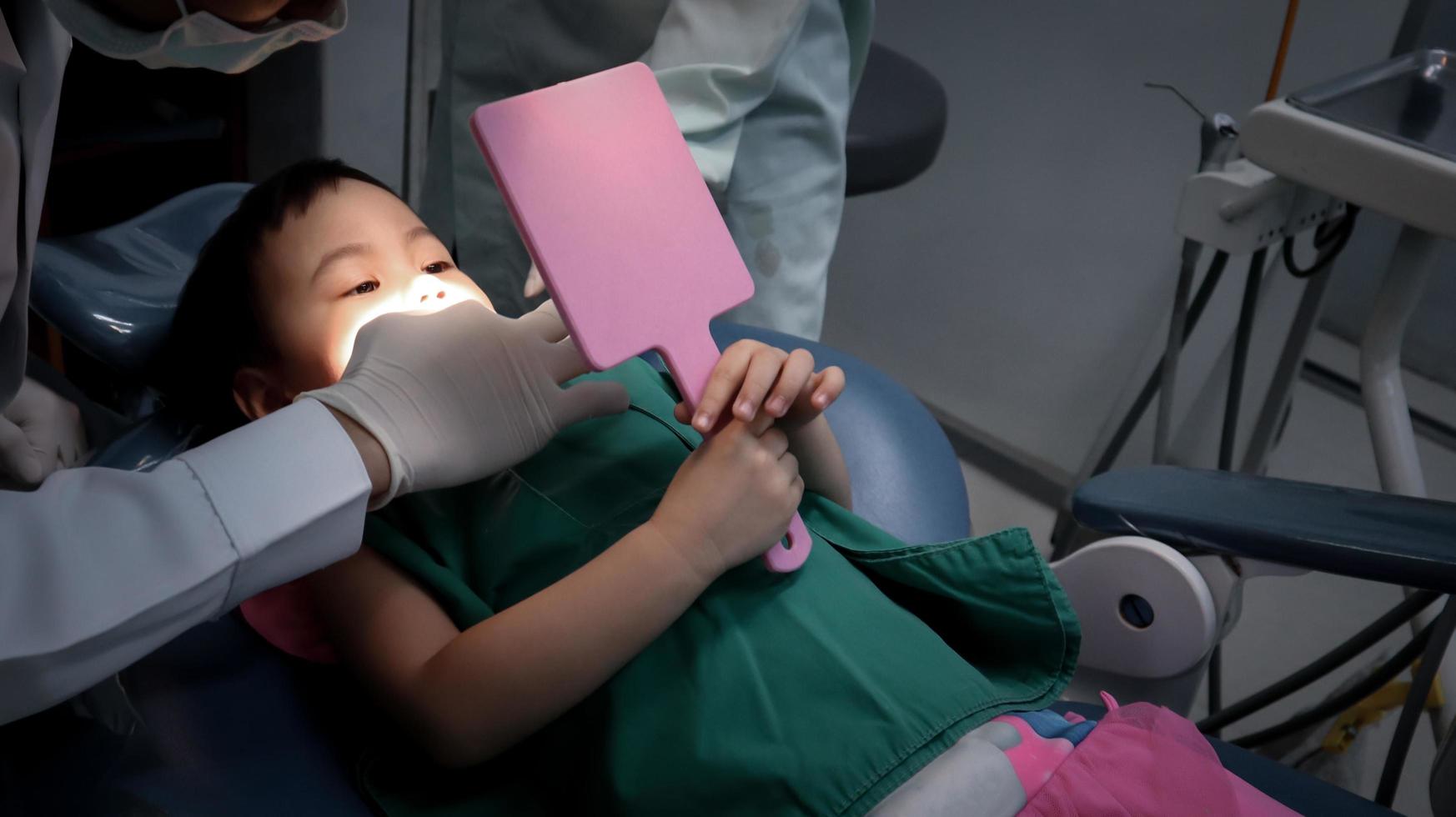 encantadora menina asiática de 3 anos na cadeira do dentista segurar o espelho para o check-up dos dentes. foto