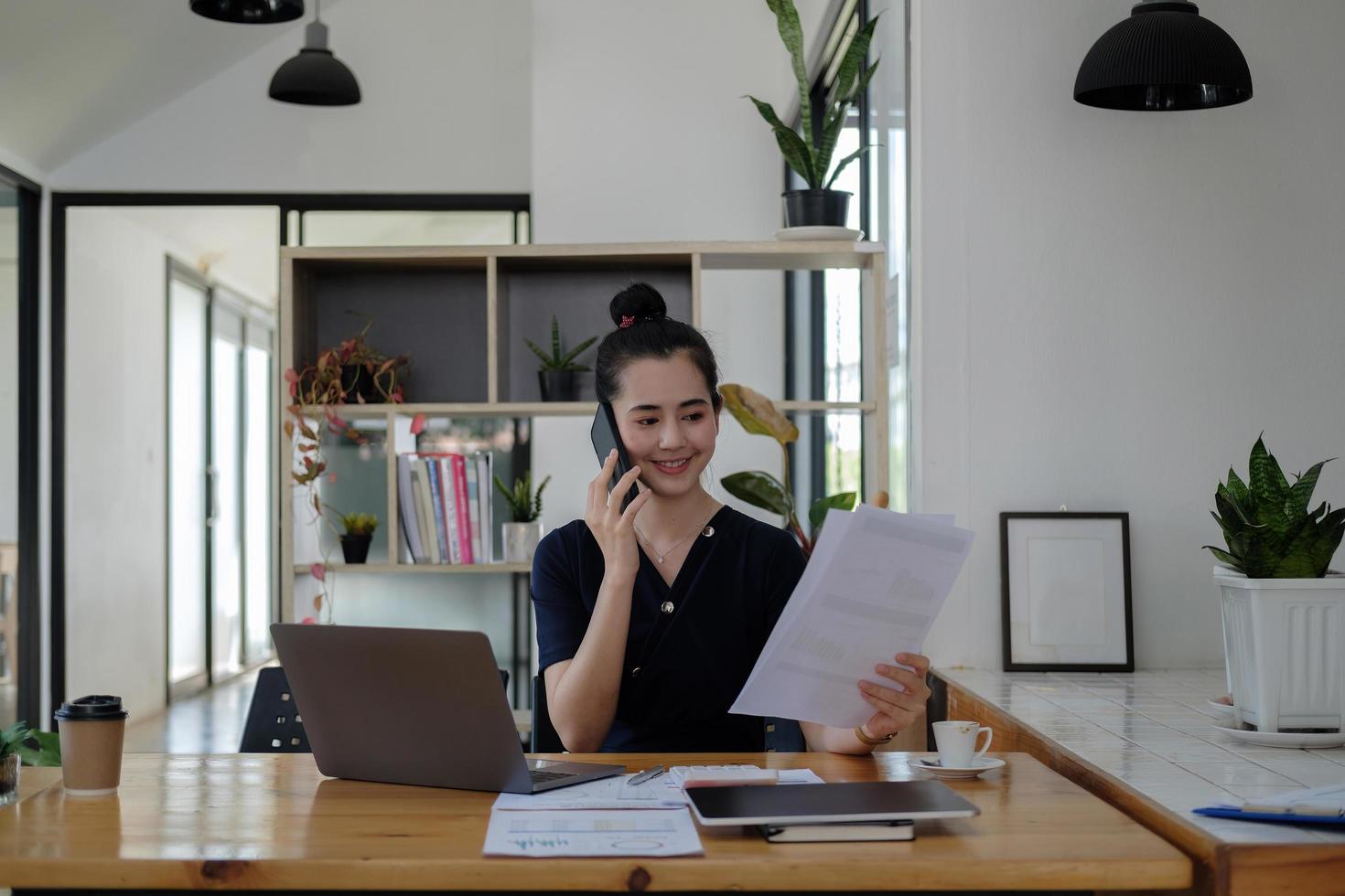 jovem empresária asiática linda encantadora sorrindo e falando no celular trabalhando com papelada gráfico gráfico financeiro no escritório foto