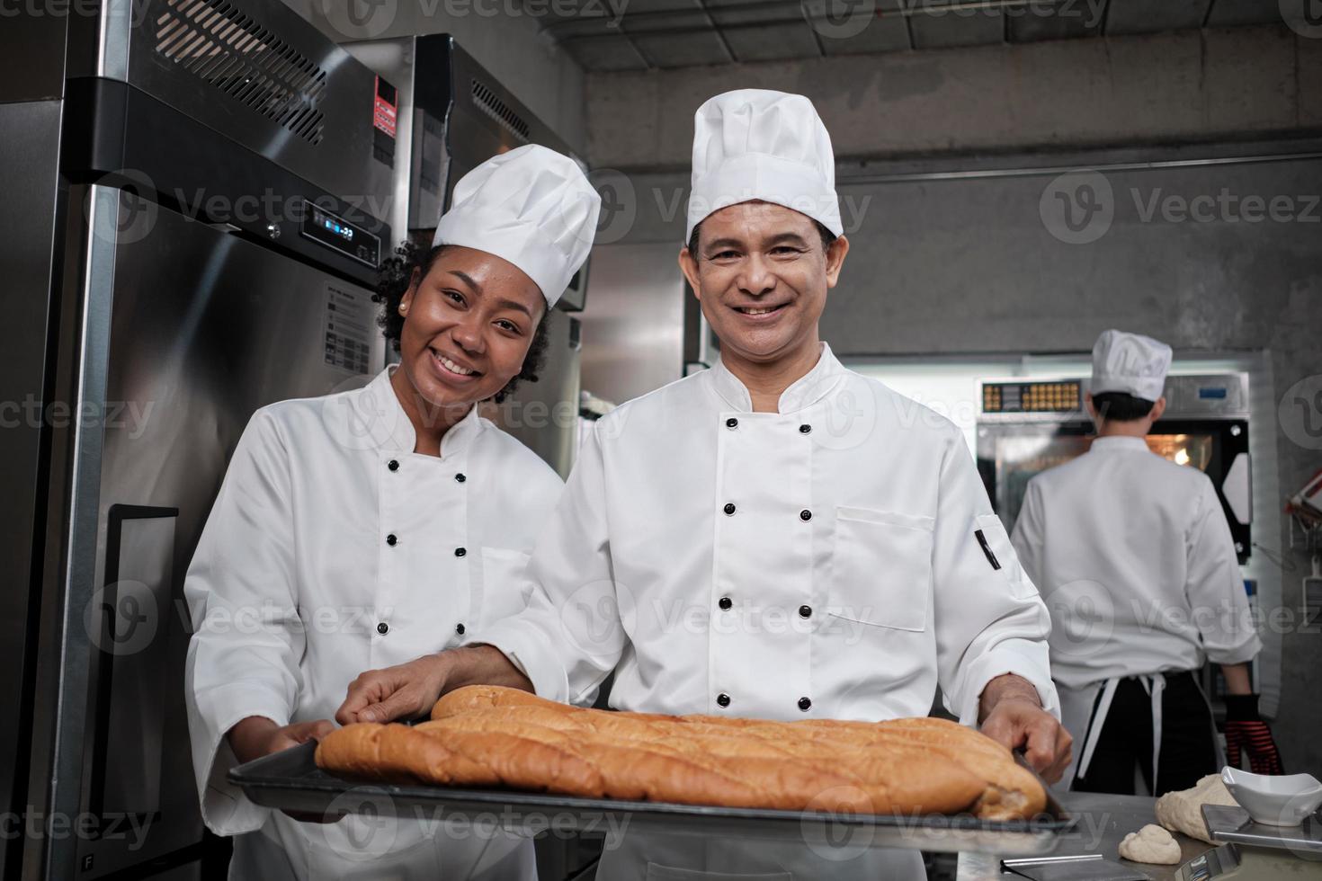 retrato de chefs profissionais de uniforme branco olhando para a câmera com um sorriso alegre e orgulhoso com bandeja de pão na cozinha. um amigo e parceiro de alimentos de pastelaria e ocupação diária de padaria fresca. foto