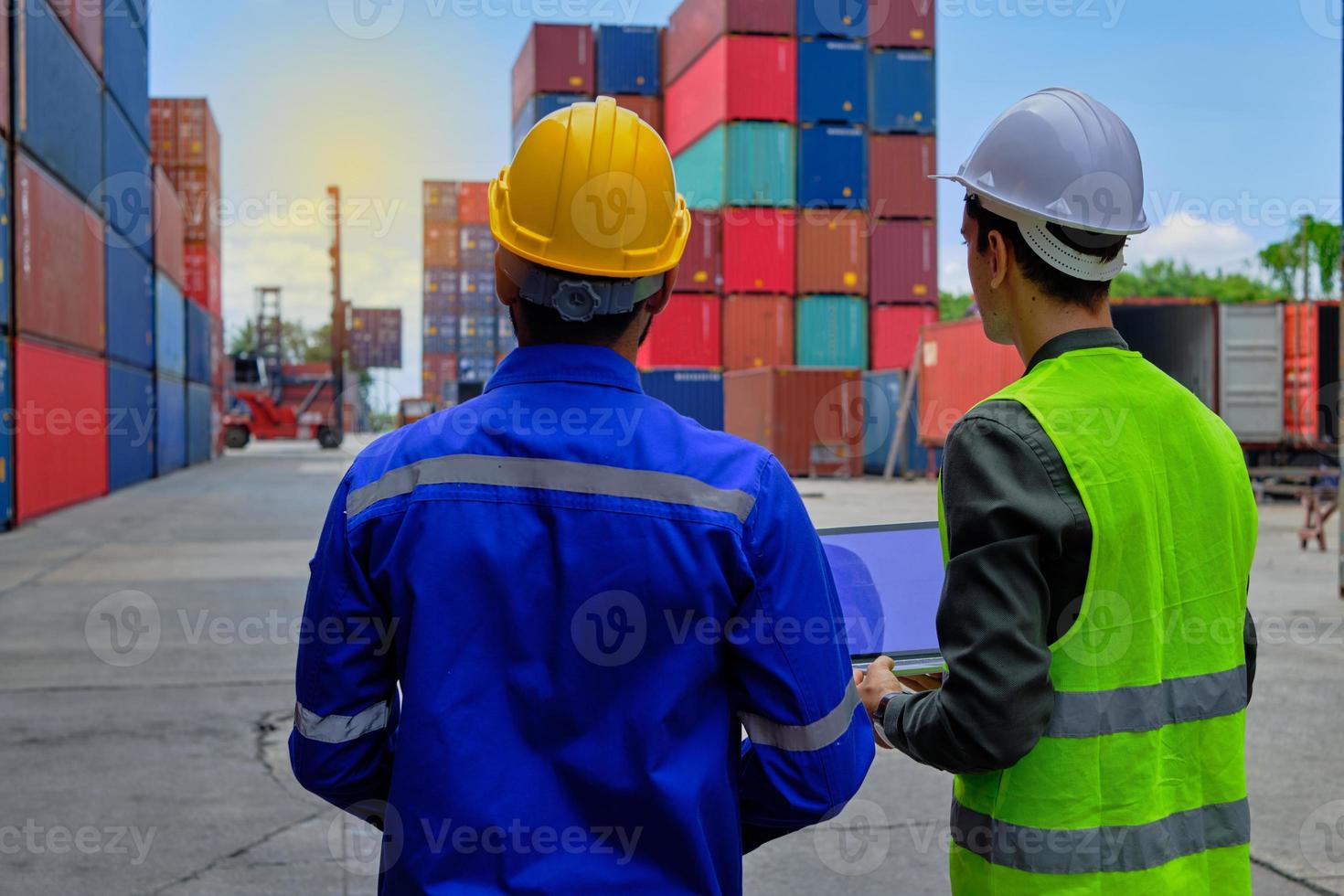 dois trabalhadores masculinos profissionais em uniformes de segurança e capacetes trabalham e inspecionam um terminal logístico com muitas pilhas de contêineres, carregando mercadorias de controle de transporte para a indústria de transporte de carga. foto