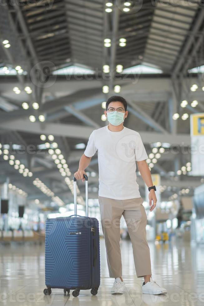 jovem homem usando máscara facial com bagagem andando no aeroporto, proteção contra infecção por doença de coronavírus, viajante asiático com chapéu. hora de viajar após o conceito de dose de reforço de vacina foto