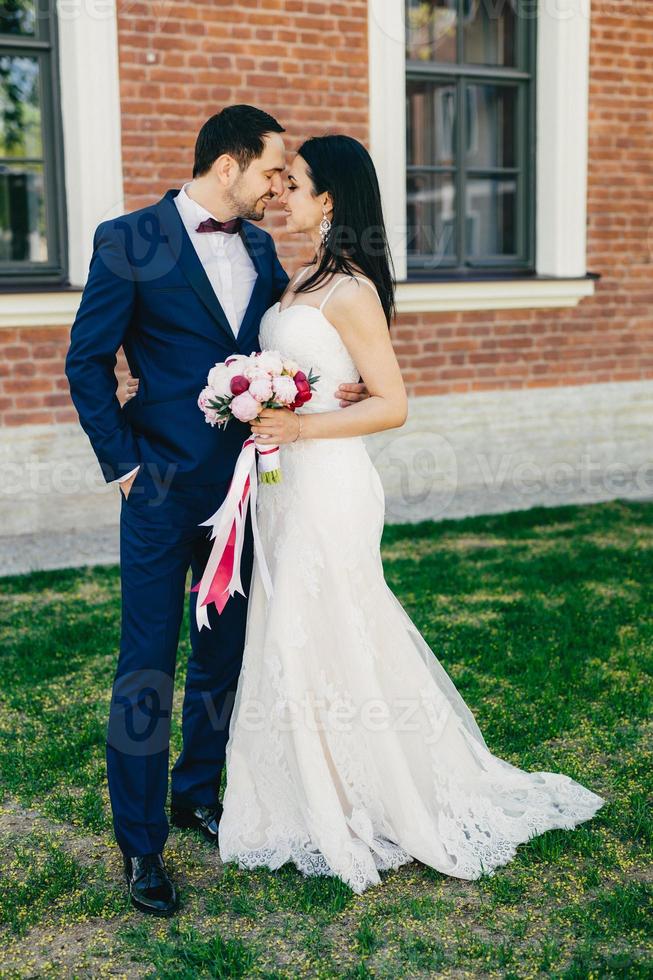 afetuoso casamento jovem casal vai se beijar, sendo feliz em celebrar seu casamento. linda mulher morena de vestido branco e noivo bonito terno preto elegante. conceito de união foto