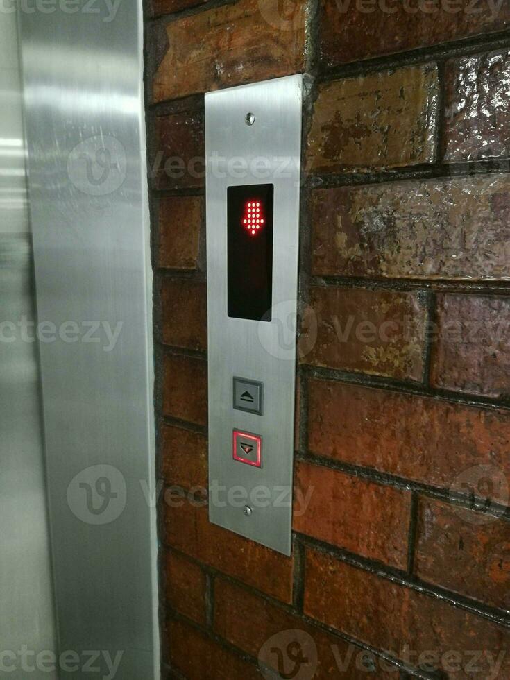 o teclado do elevador tem uma luz vermelha no botão para baixo e um símbolo de seta para baixo. teclado está montado ao lado do elevador em uma parede marrom. foto