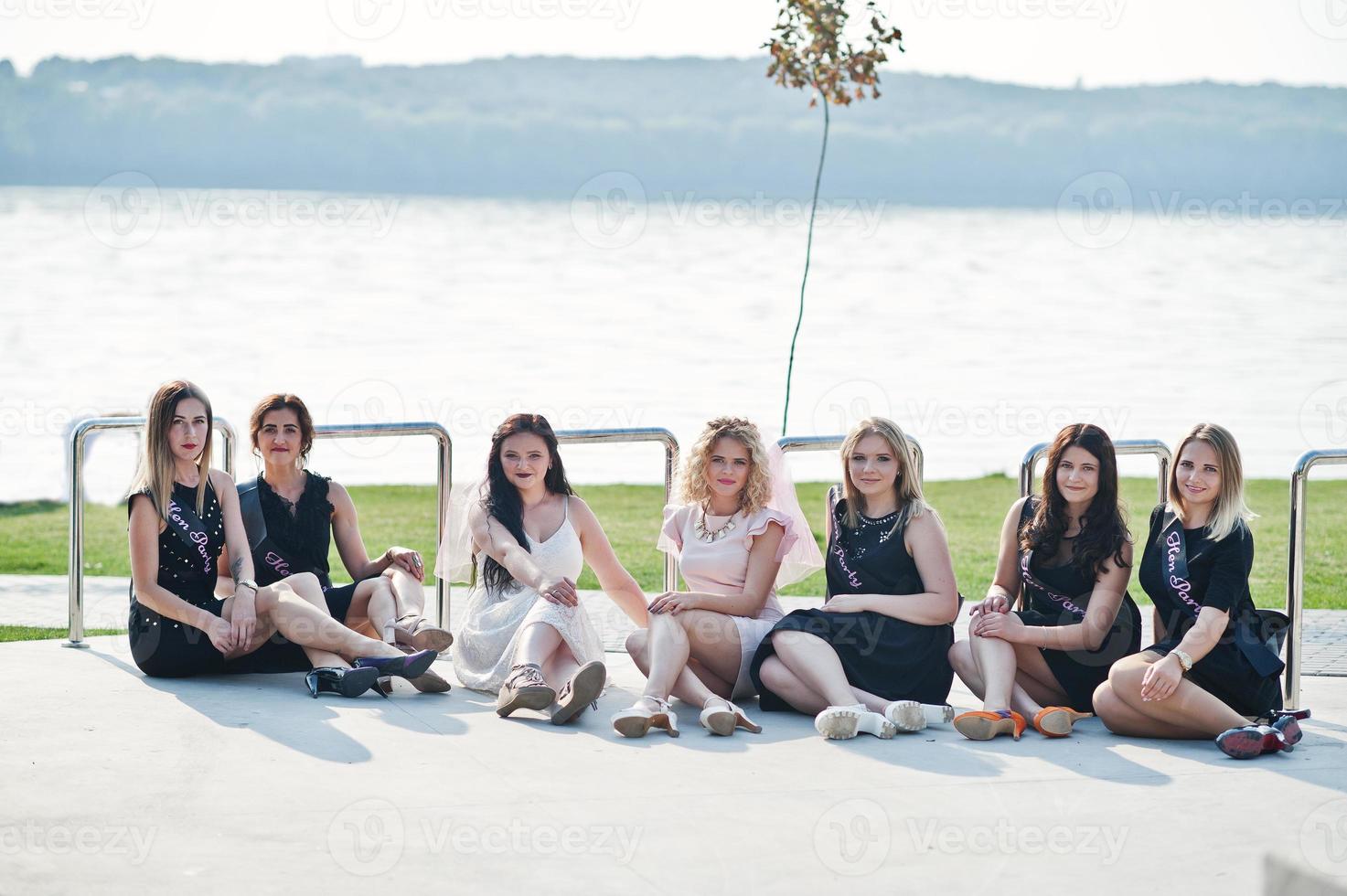 grupo de 7 meninas usam preto e 2 noivas sentadas no lago de fundo na festa de despedida de solteira. foto