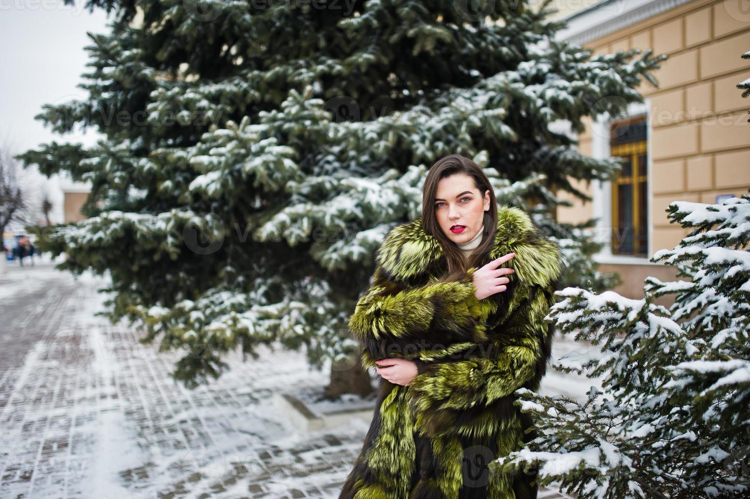 menina morena com casaco de pele verde em dia de inverno contra pinheiro nevado. foto