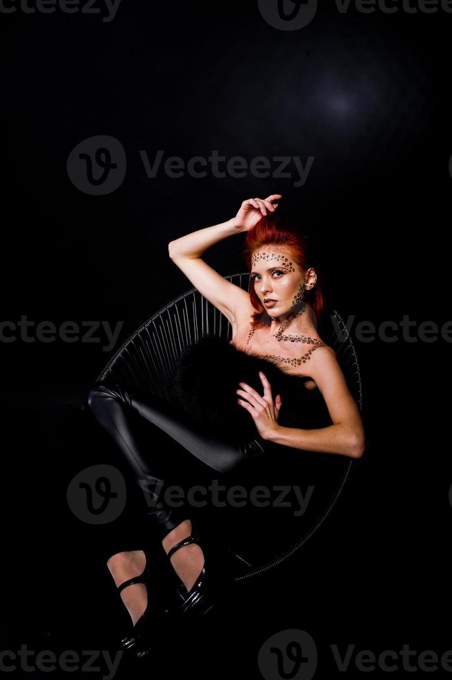 menina ruiva modelo de moda com maquiagem originalmente como predador de leopardo isolado em preto. retrato de estúdio na cadeira. foto