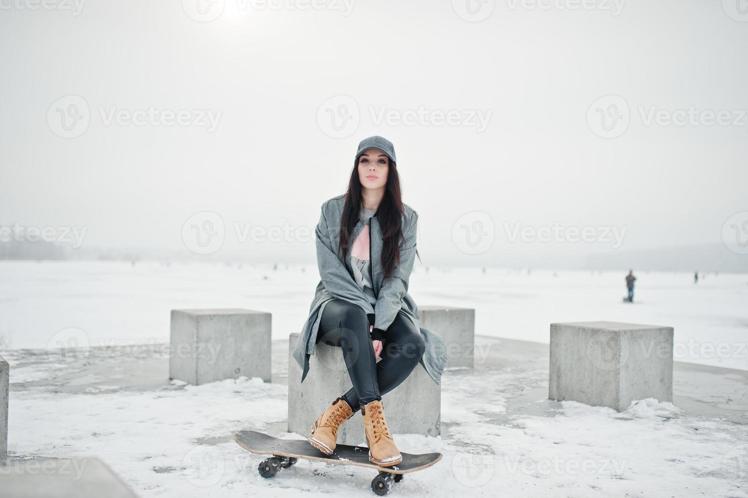 menina morena elegante boné cinza, estilo casual de rua com skate em dia de inverno. foto