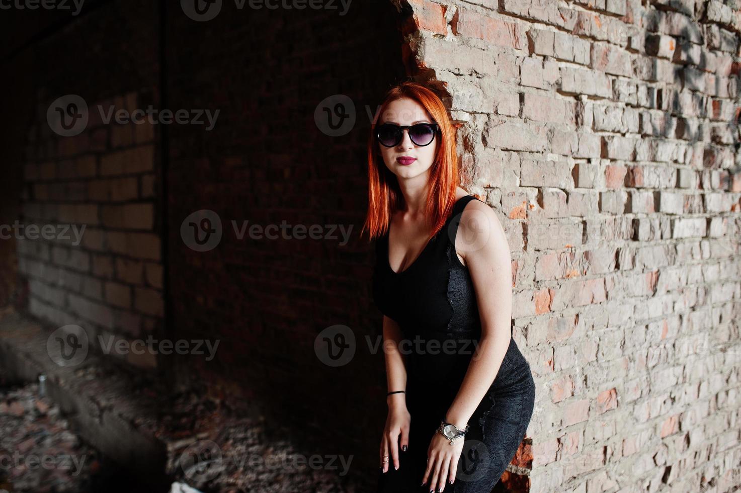 garota elegante ruiva em óculos de sol usam preto, contra um lugar abandonado com paredes de tijolo. foto